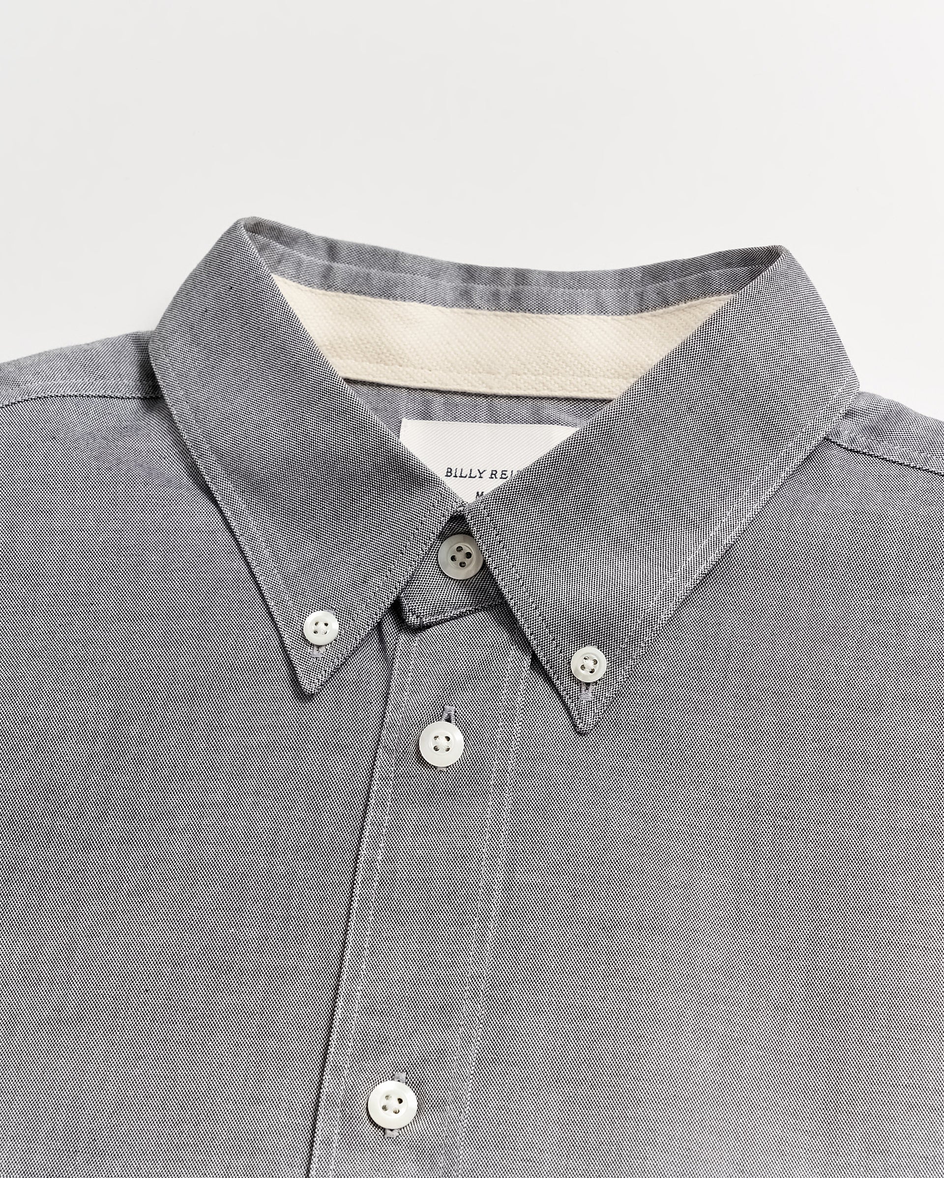 A close-up of a grey button-down shirt with a pointed collar and white buttons, displayed on a flat white surface. A label is partially visible inside the collar.
