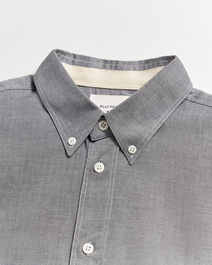 A close-up of a grey button-down shirt with a pointed collar and white buttons, displayed on a flat white surface. A label is partially visible inside the collar.