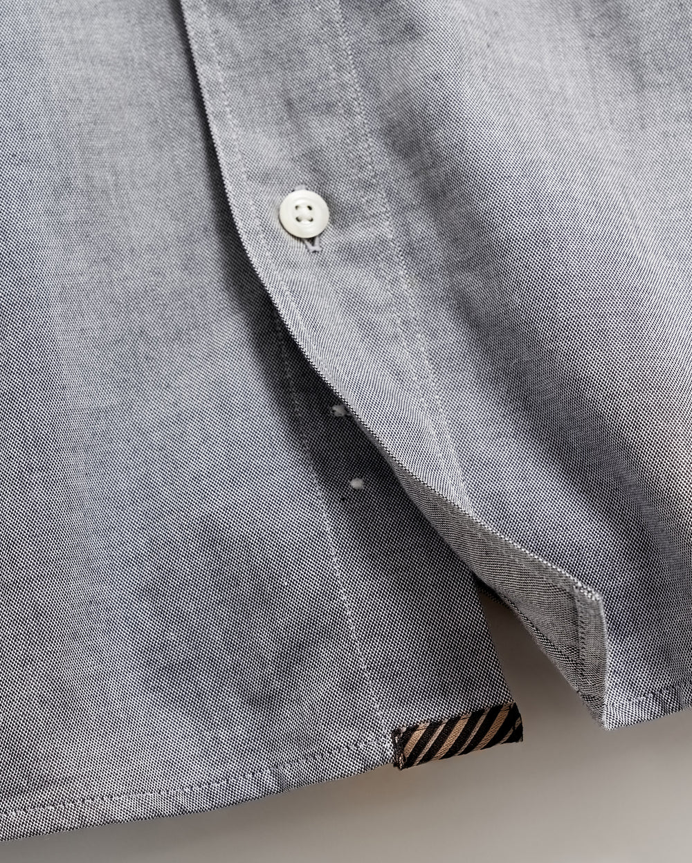 Close-up of a light gray shirt with a white button partially fastened, revealing the inside seam and a small black and brown striped fabric detail at the hem.