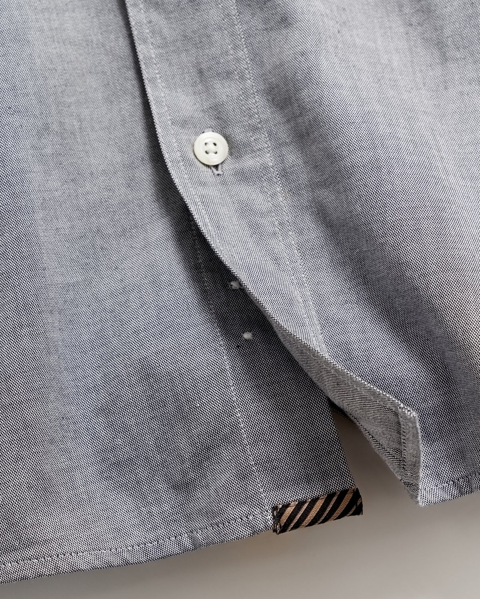 Close-up of a light gray shirt with a white button partially fastened, revealing the inside seam and a small black and brown striped fabric detail at the hem.