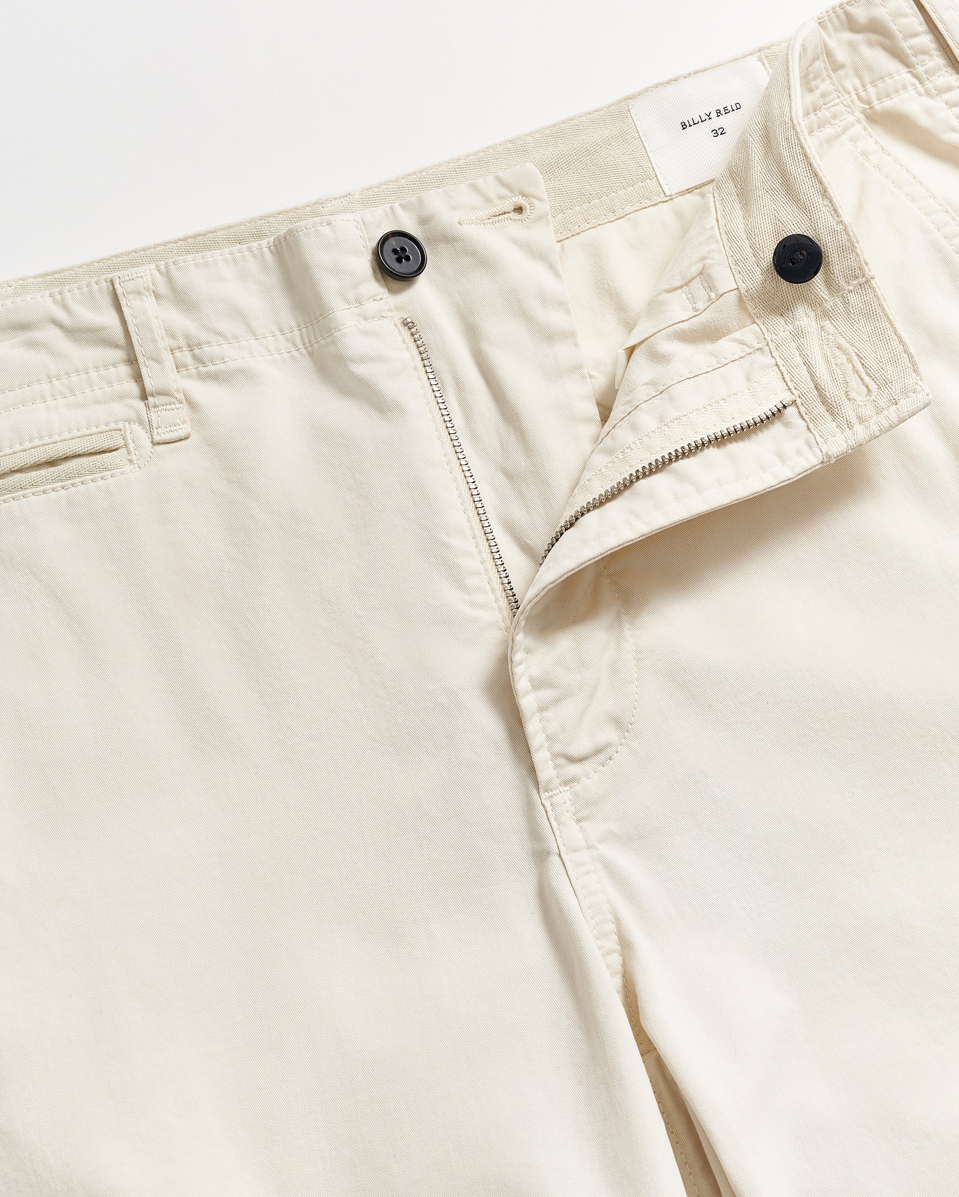 A close-up of cream-colored pants with the fly partially unzipped, showing a button closure and waistband label that reads BILLY REID 33. The pants feature belt loops and slanted front pockets.