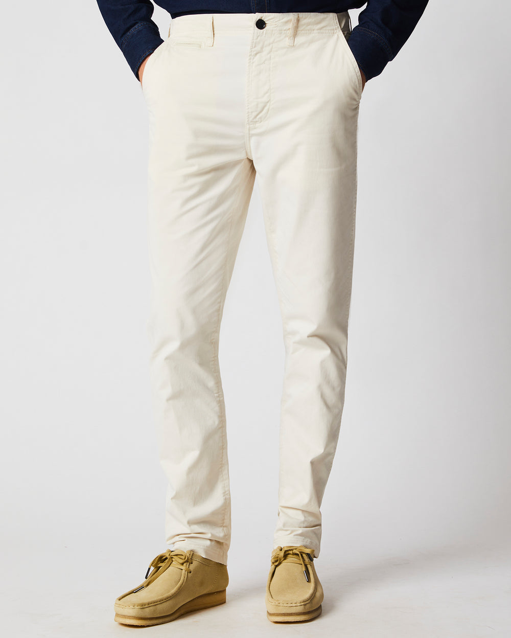 A person wearing off-white pants and tan suede shoes stands against a plain light background, with their hands in their pockets. Their upper body is partially visible, dressed in a dark long-sleeve shirt.