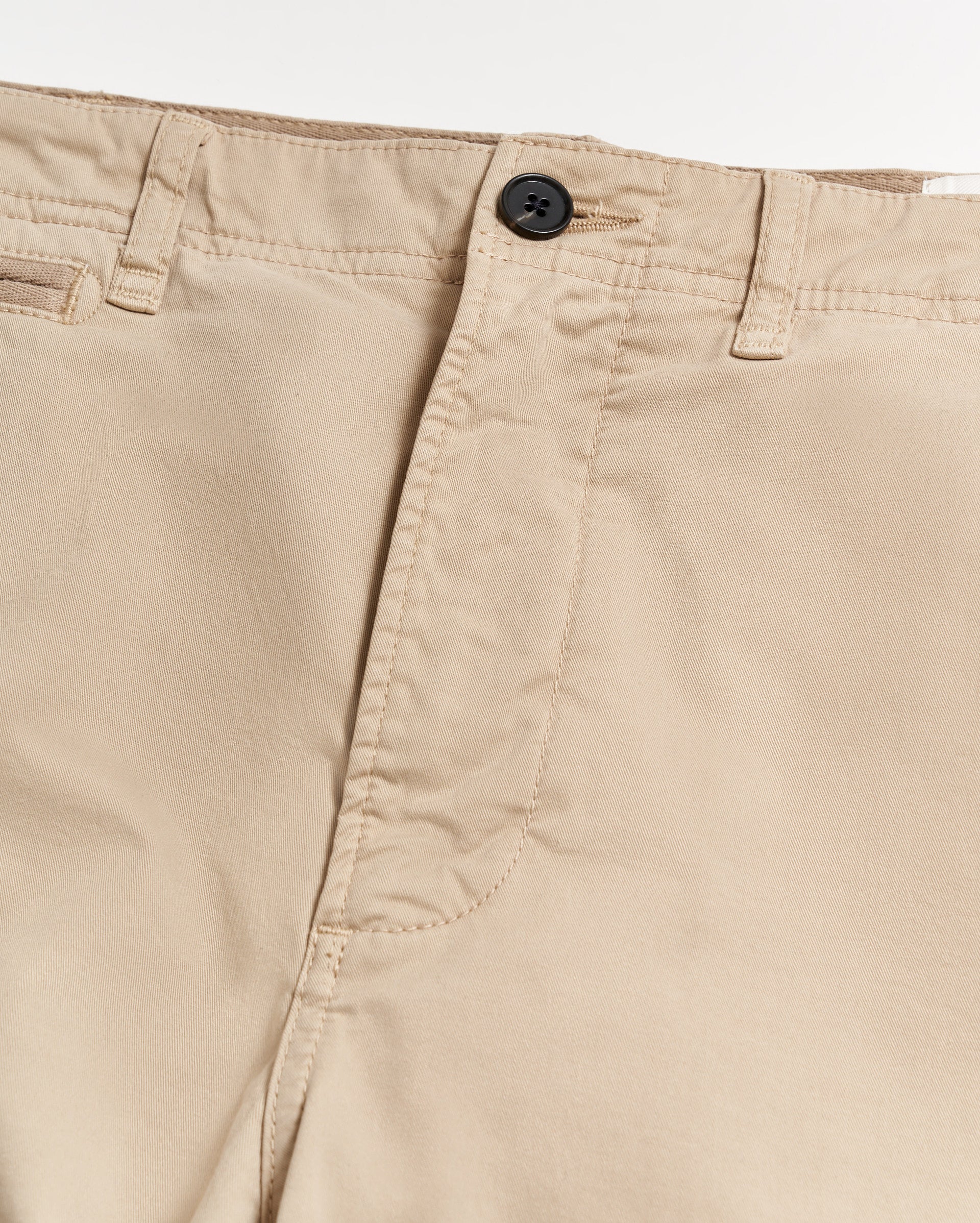 Close-up of the waistband and upper front of beige pants, showing a black button, zipper fly, belt loops, and stitching details on the light fabric.