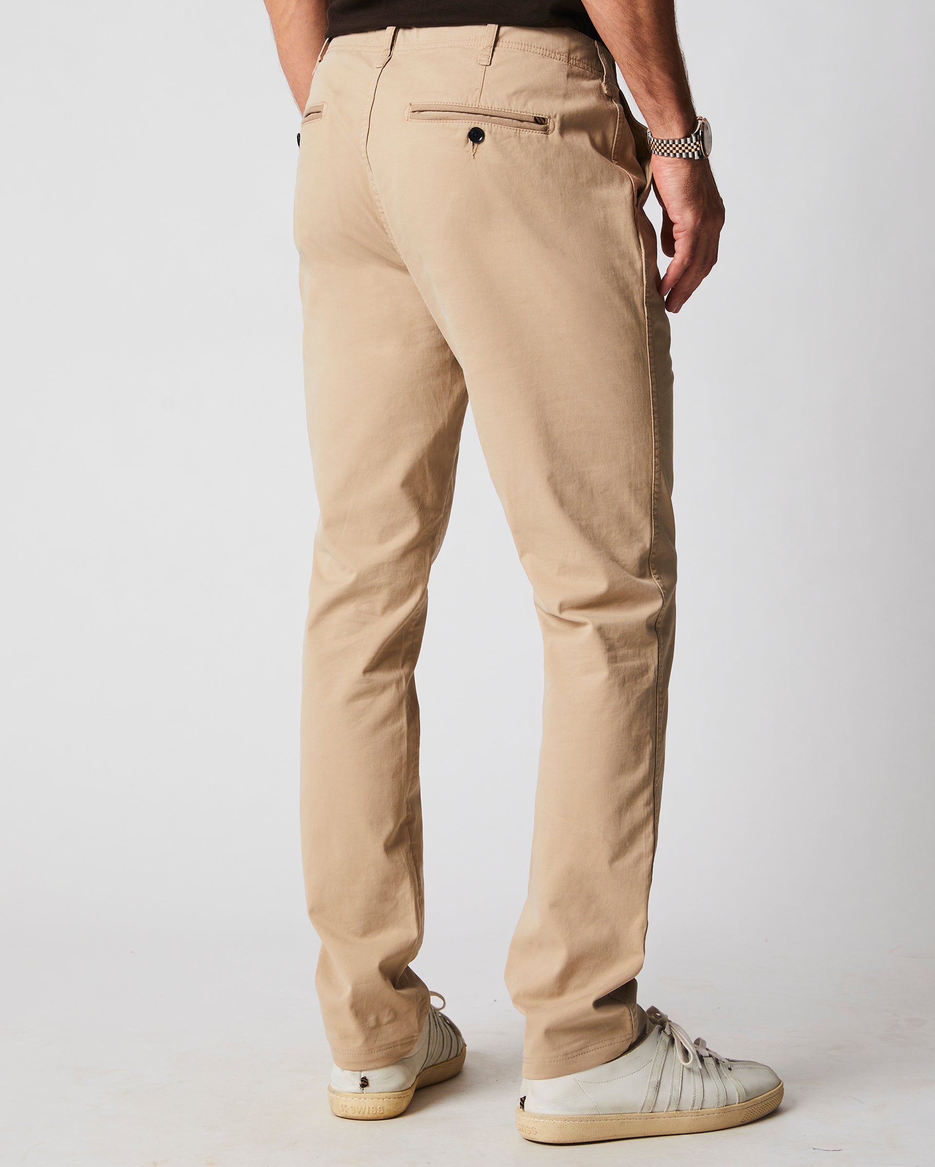 A man is standing facing away, wearing beige chinos, a black shirt, a silver watch, and white sneakers against a plain light background.