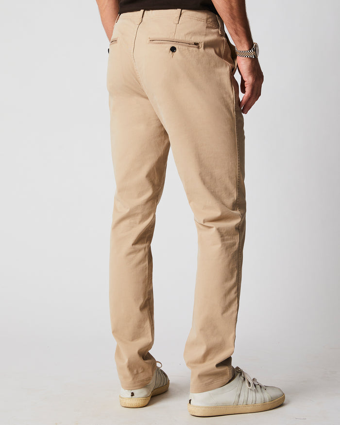 A man is standing facing away, wearing beige chinos, a black shirt, a silver watch, and white sneakers against a plain light background.