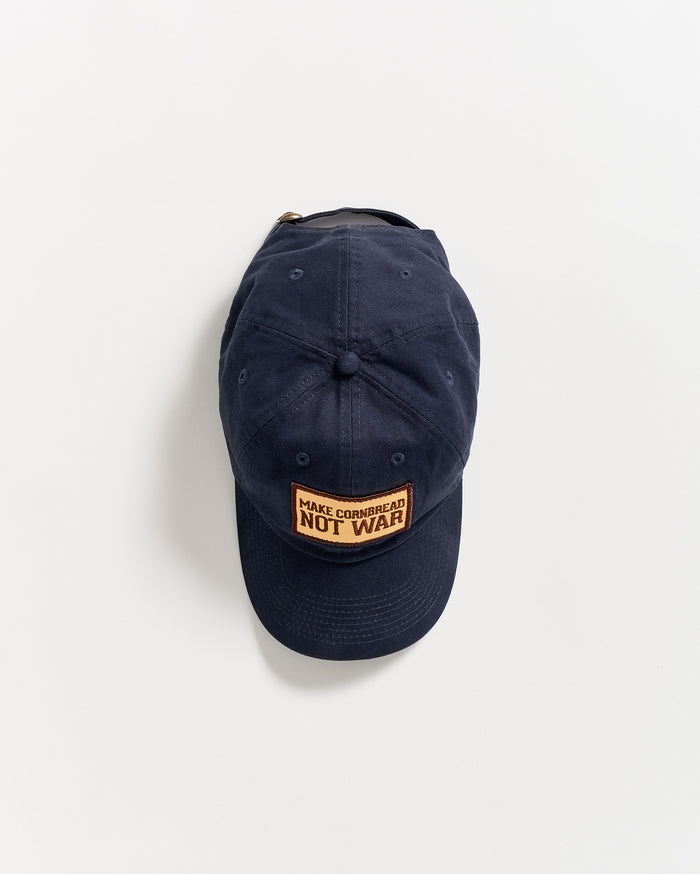 A navy blue baseball cap with a rectangular patch on the front that reads MAKE CORNBREAD NOT WAR in orange and black text, photographed against a plain white background.
